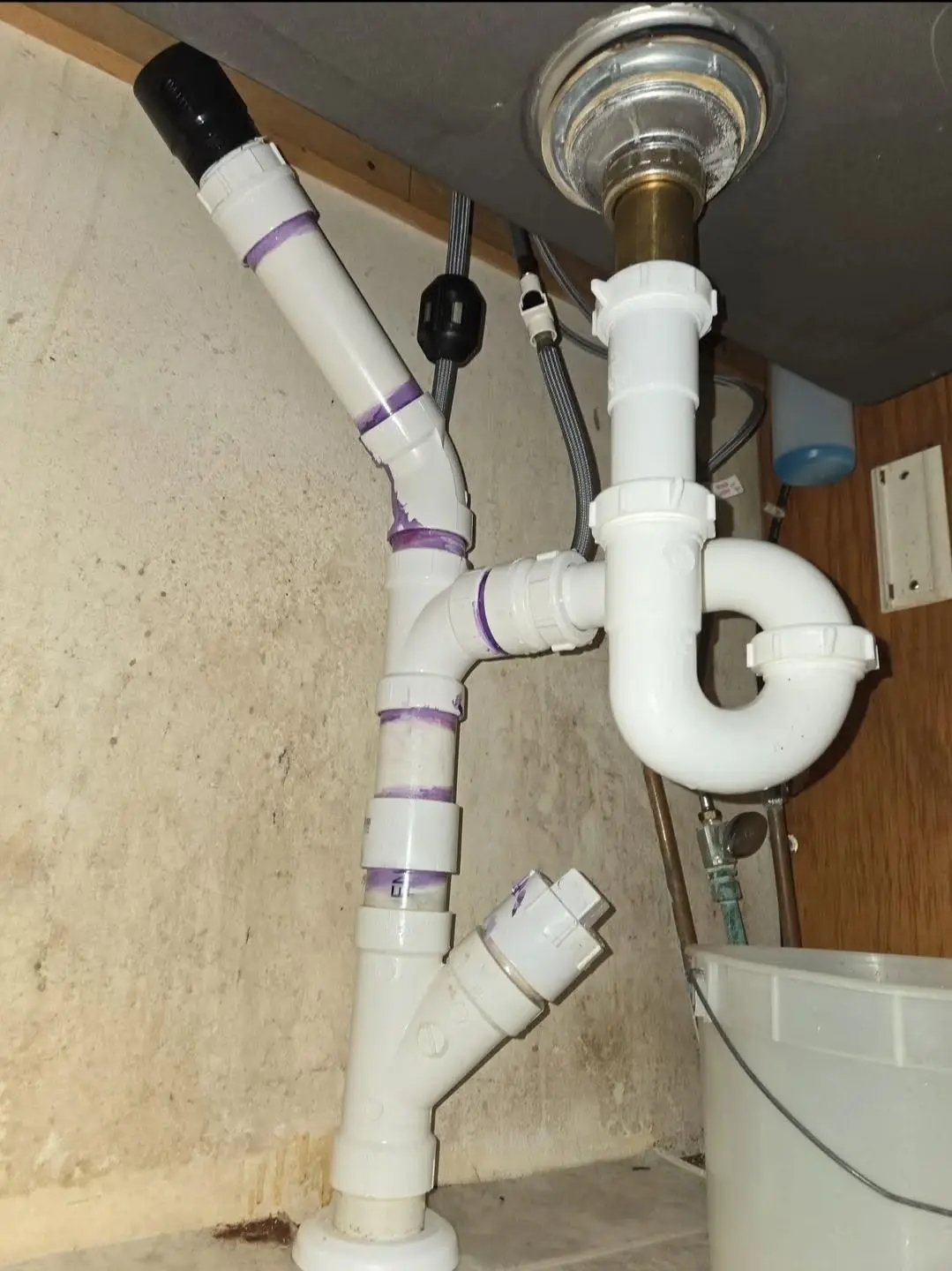 Rebuilt white plastic drain under a kitchen sink with a wye fitting and a cleanout plug, primer marks on the joints